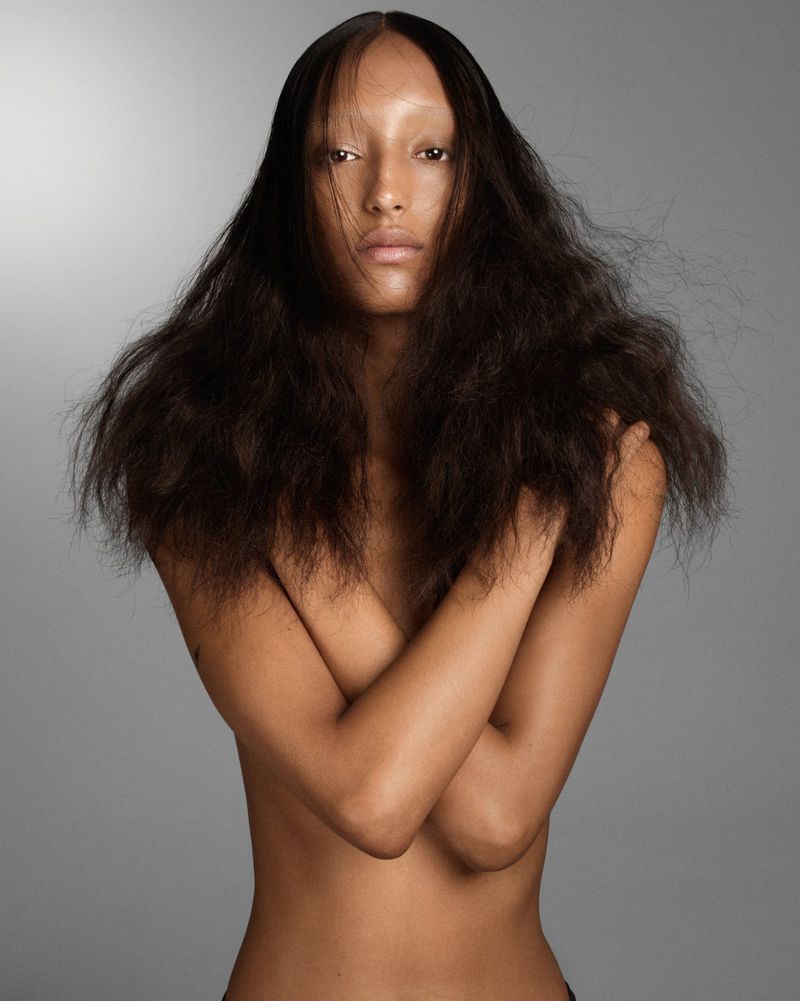 Striking fashion portrait of a bare-chested biracial adult female model with textured dark hair against a solid grey backdrop.