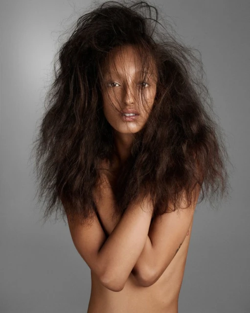 Striking studio portrait of an adult woman with long, dark, tousled hair and tanned skin, posed against a plain gray backdrop.