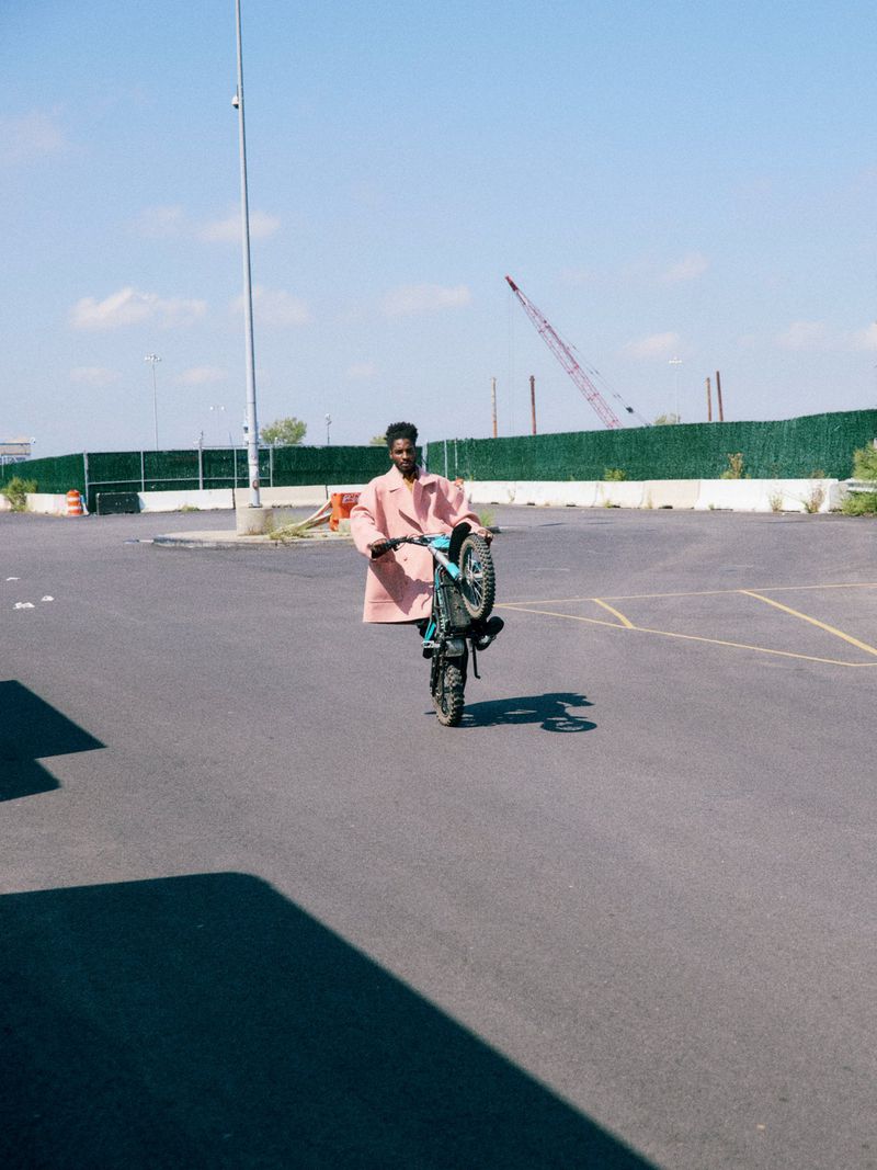 An African American man on an electric bicycle doing a wheelie in an empty parking lot on a sunny day, wearing a pink coat.