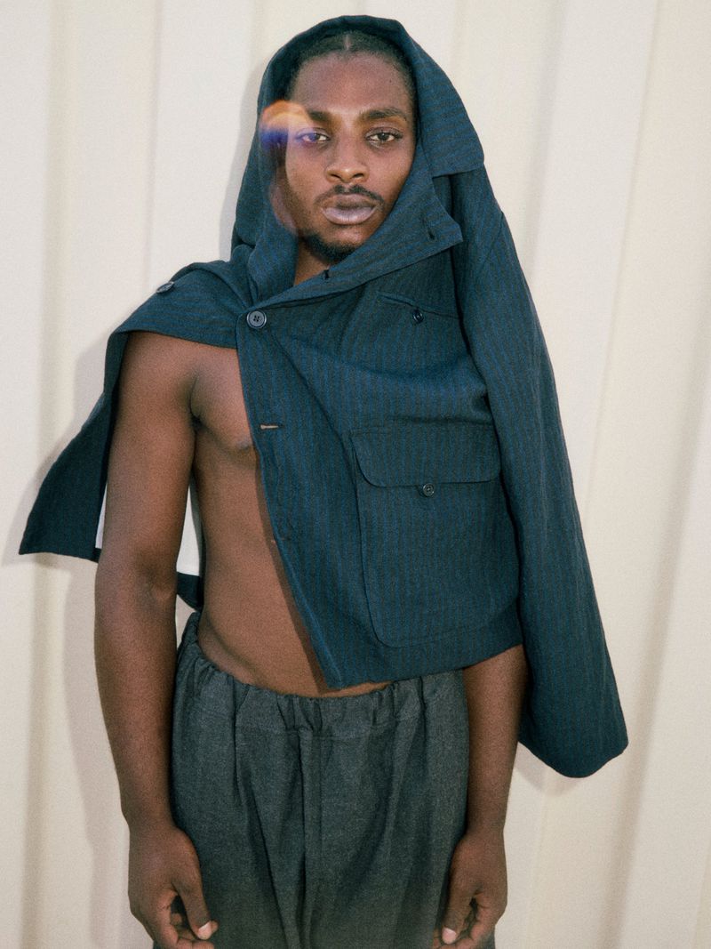 Portrait of an African American man with a jacket draped over his head and shoulders, wearing gray pants, posed against a light background.
