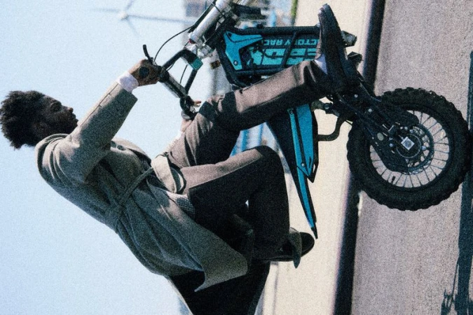 Adult male in a trench coat riding an electric dirt bike on an urban street, photographed at an extreme angle against a blue sky.