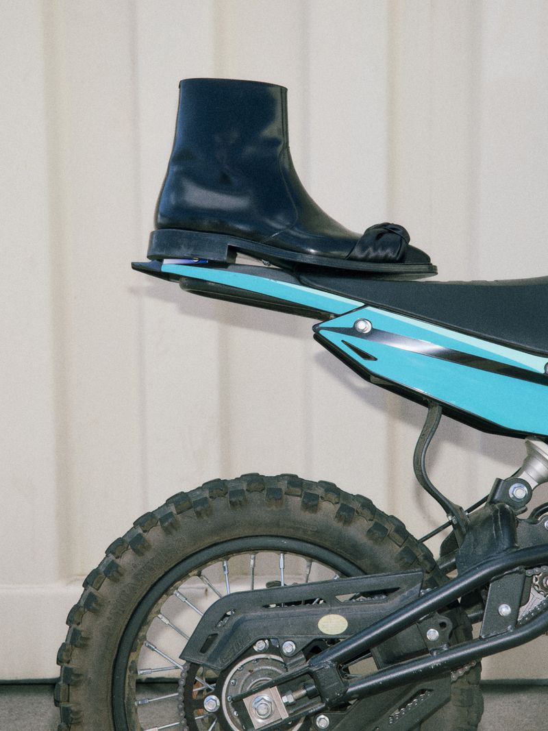 Fashionable black leather boot rests on a teal dirt bike against a clean white backdrop, showcasing style and utility.