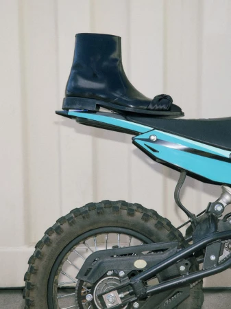 Fashionable black leather boot rests on a teal dirt bike against a clean white backdrop, showcasing style and utility.