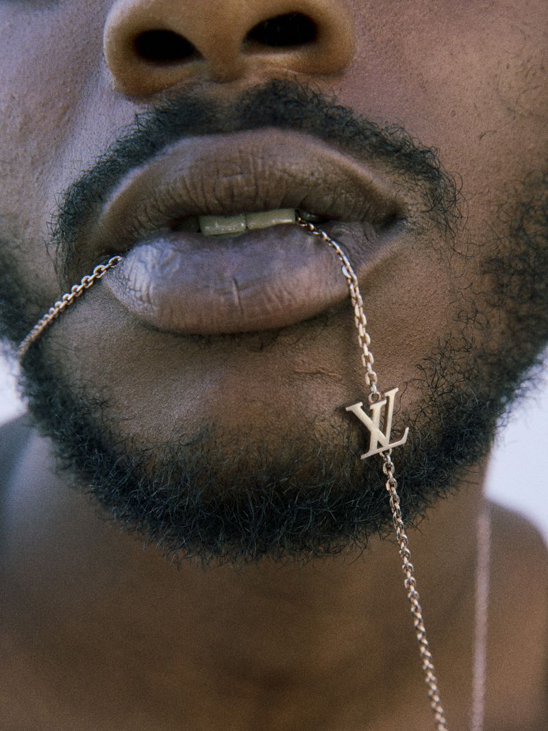Close-up of a person with dark skin and facial hair, wearing a designer chain that goes in the mouth, with bright lighting.