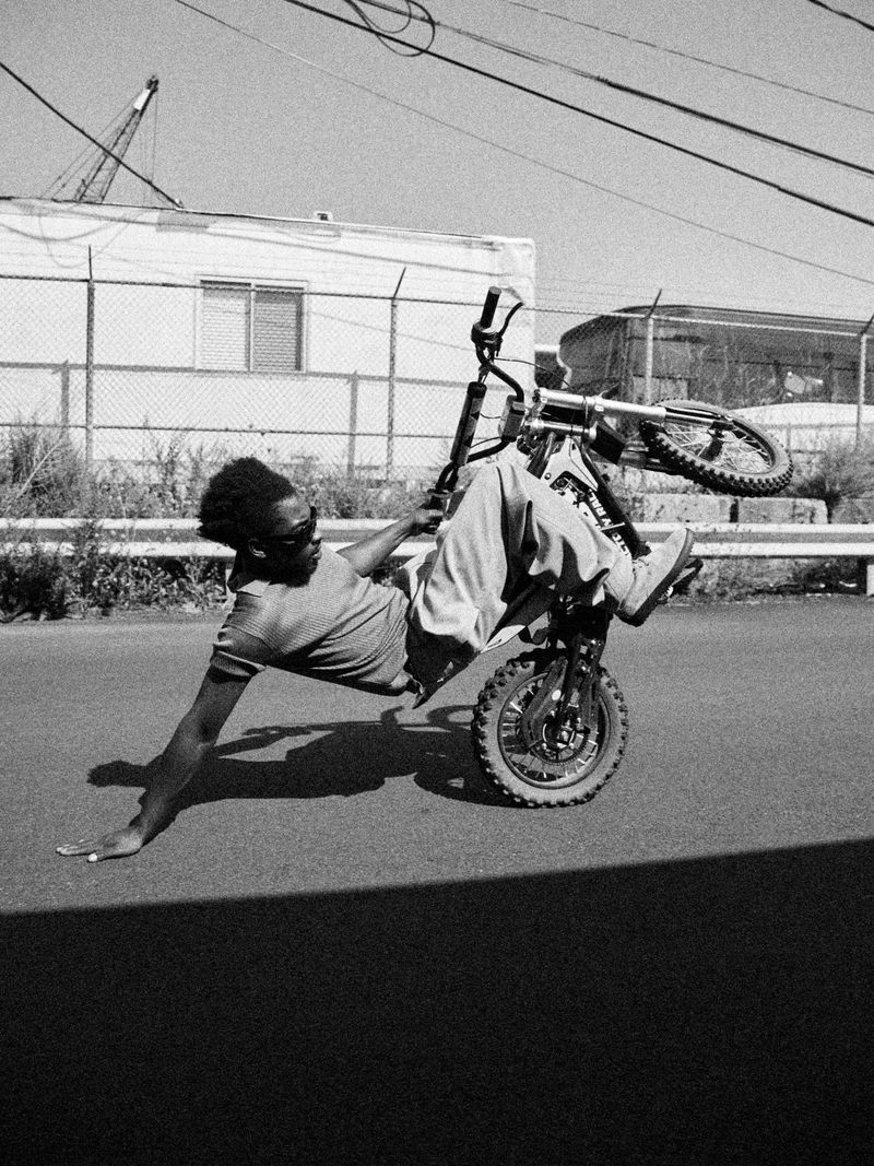 A man balances on a dirt bike on the front tire while lying sideways on the seat in a black and white outdoor shot.