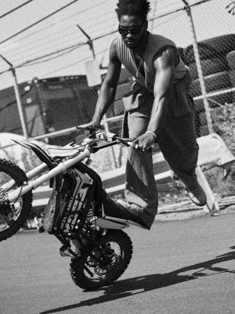 Monochrome portrait of a man performing a wheelie on a dirt bike on a paved road with a chain link fence in the background.