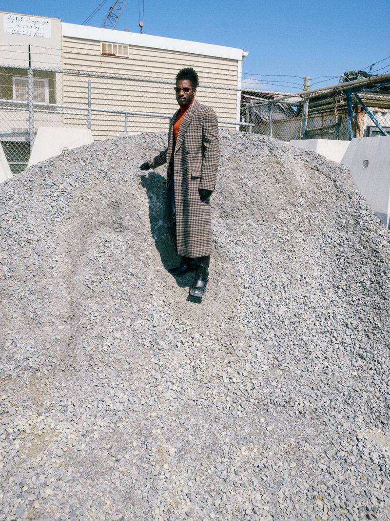 Adult male in plaid coat stands atop gravel hill outside on sunny day; urban background, clear blue sky.