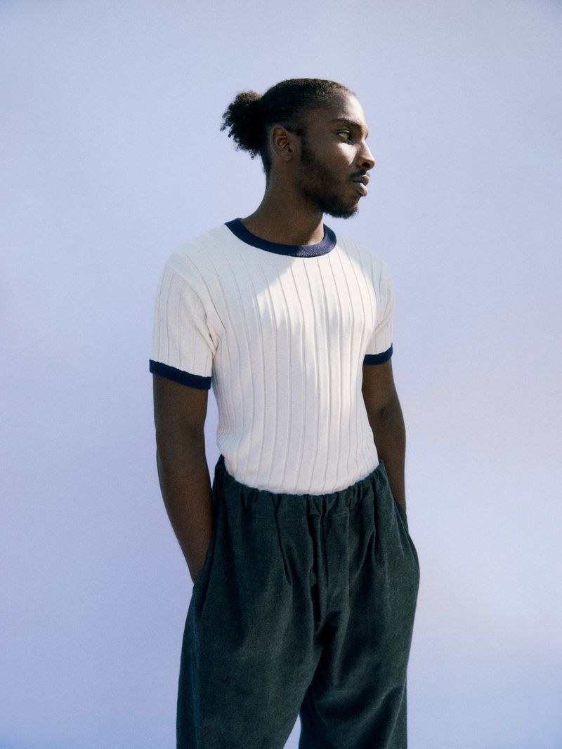 Portrait of an adult Black male wearing a ribbed white shirt and dark pants, looking to the side against a light blue background.