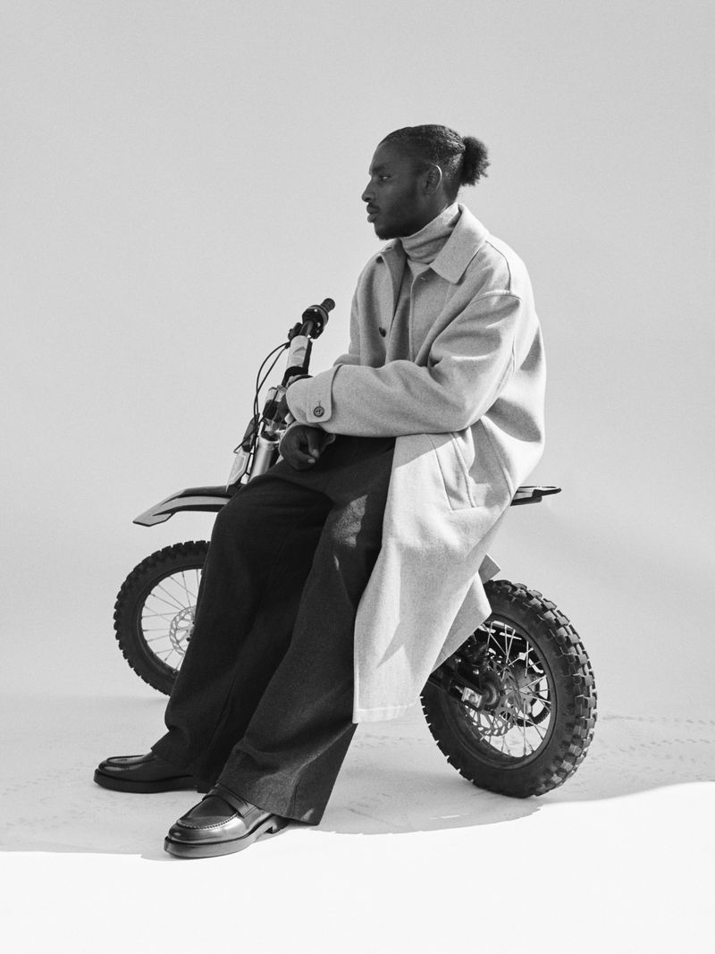 Black man in a grey coat sits on a dirt bike in a photo studio with a neutral background and relaxed posture, dressed in stylish black pants and loafers.