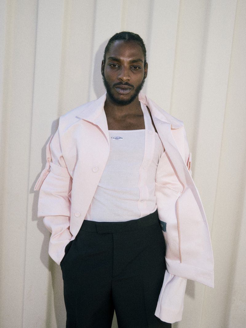 Studio portrait of an African American man in a pink jacket, white tank top, and black pants, posing against a cream curtain.