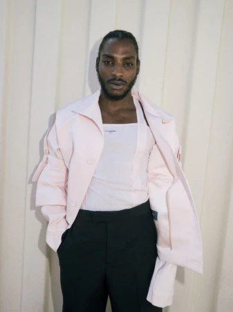 Studio portrait of an African American man in a pink jacket, white tank top, and black pants, posing against a cream curtain.