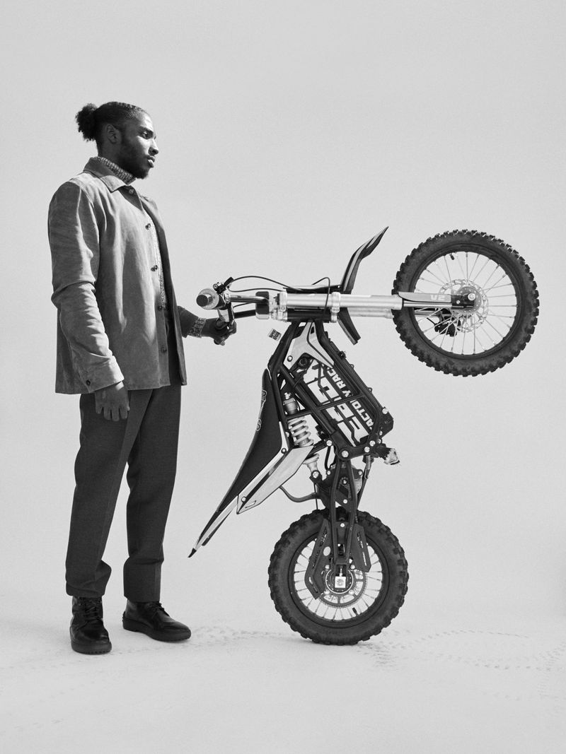 An adult male in a jacket and pants stands next to a dirt bike, holding it up by the handlebars in a studio with neutral lighting.