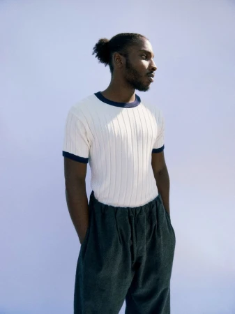 Black man with hair in a bun wearing a white ringer t-shirt and dark trousers against a pale blue background