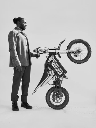 A man in a grey jacket and trousers stands in a studio holding an electric dirt bike upright on its rear wheel against a white background.