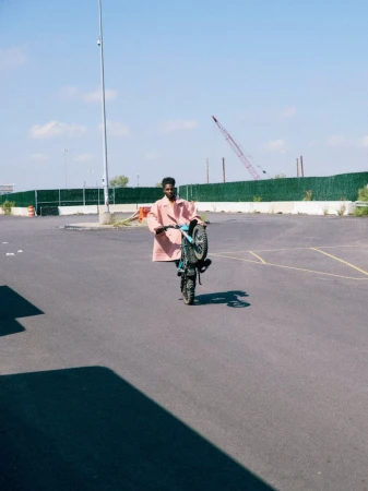 Man wearing a pink coat riding a dirt bike on one wheel in an empty asphalt lot under a clear sky.