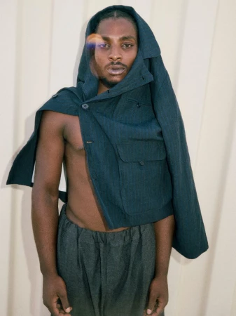 Black male model with a dark teal tailored jacket draped over his head and bare chest against a ribbed neutral backdrop.