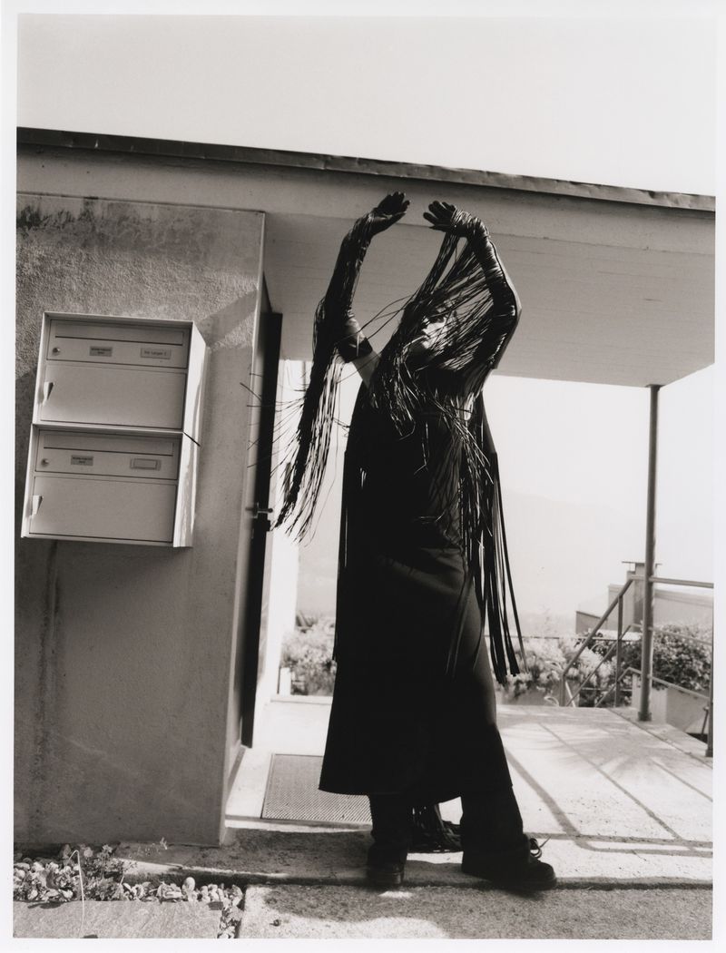 Monochrome photo of a person in a fringed garment raising their arms on a patio with a building and landscape in the background.