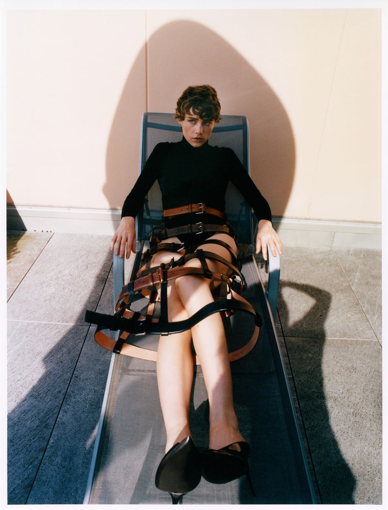 Fashionable woman in black bodysuit and belts reclining in chair in outdoor setting with neutral wall and strong shadow behind.