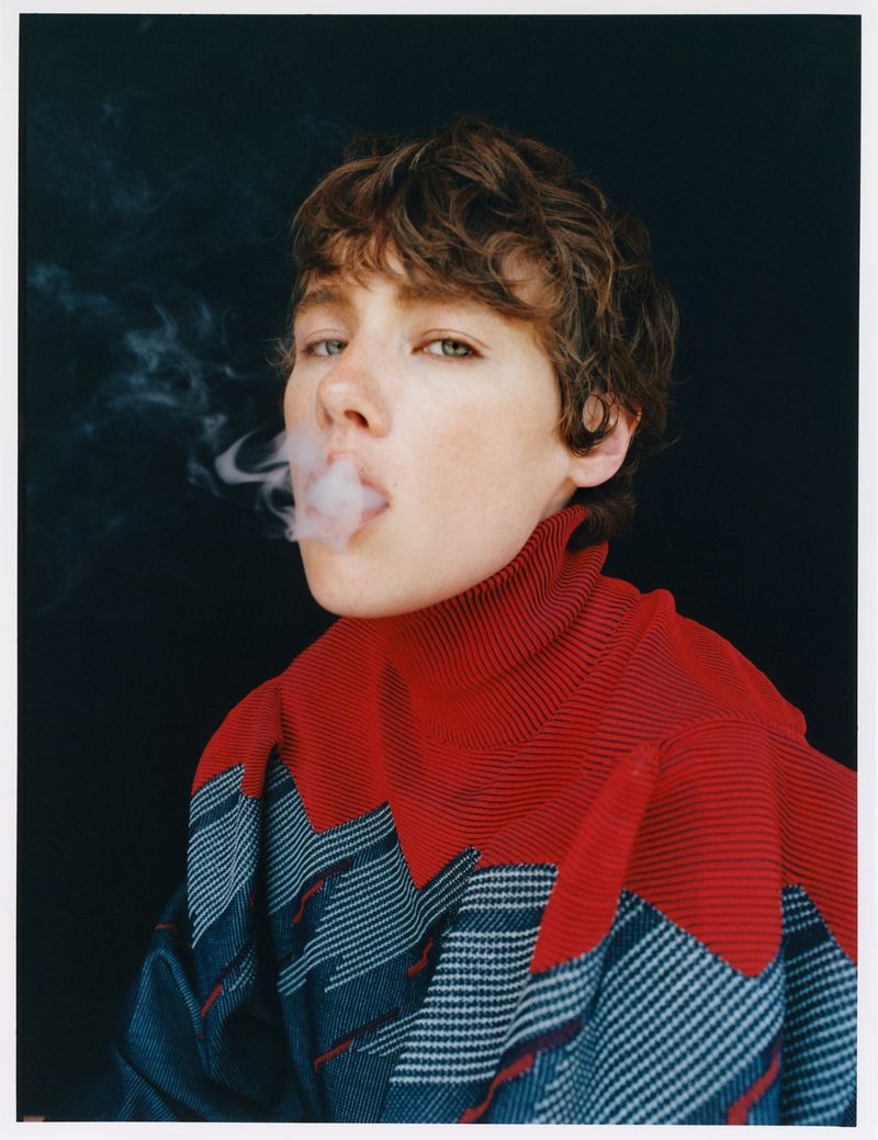 Close-up of a young adult with short brown hair blowing smoke, wearing a red and blue top against a dark background in a studio.