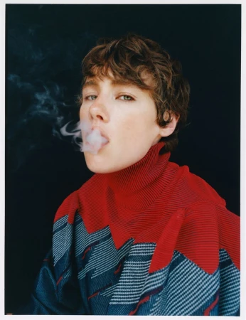 Close-up of a young adult with short brown hair blowing smoke, wearing a red and blue top against a dark background in a studio.