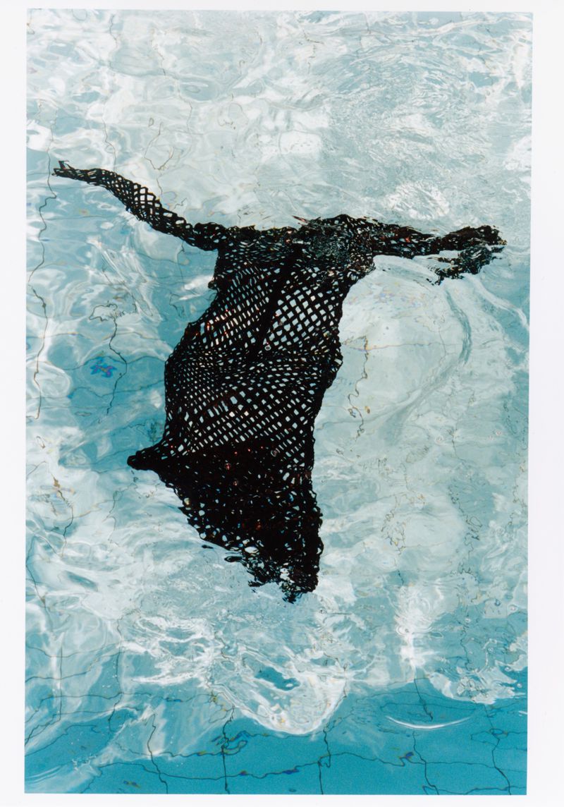 Black mesh fabric floats in clear blue pool water, creating an abstract underwater effect with sunlight patterns visible on the pool bottom.