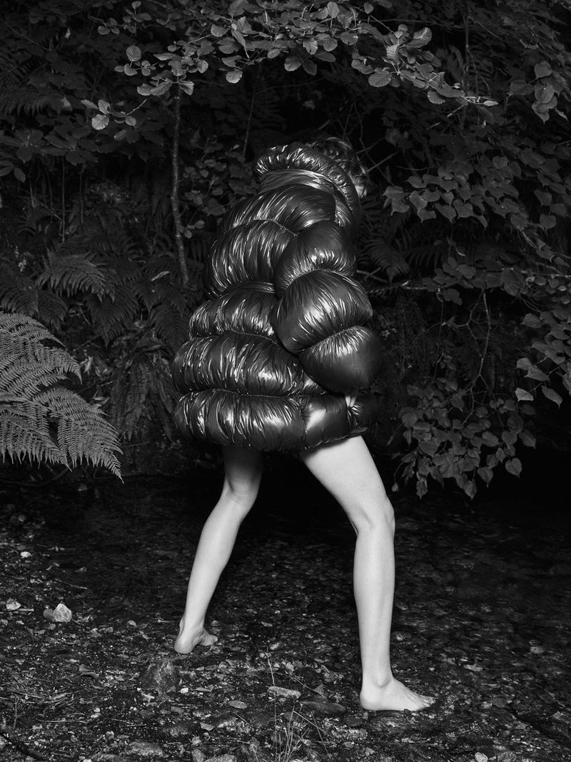 Woman in oversized puffer jacket walking barefoot on forest floor, surrounded by ferns and foliage in black and white.
