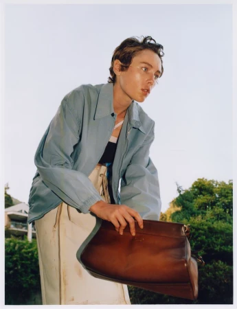 Adult female model poses outdoors, holding a large brown leather handbag, wearing a light blue jacket, beige pants against sky background.