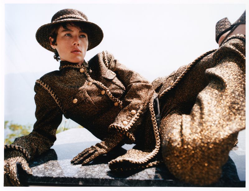 A woman in a tweed suit and hat reclines on a stone surface in bright sunlight, showcasing classic style and high-fashion design.