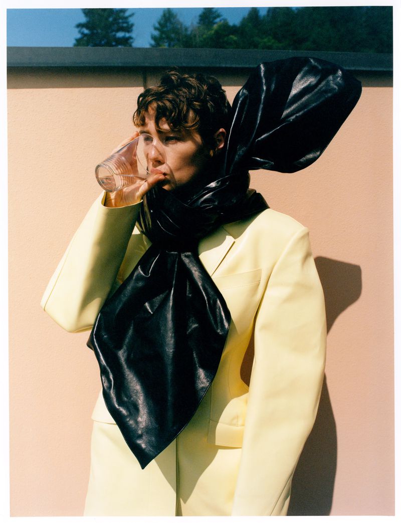 Woman in a yellow suit drinks water outdoors, wearing a large black scarf against a peach colored wall and greenery in the background.