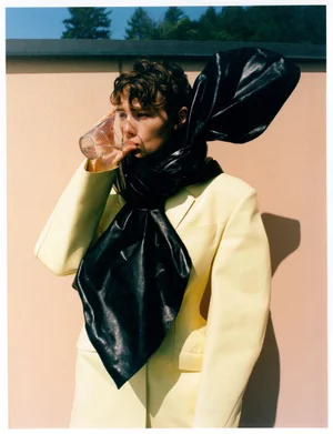 Model Annabelle Weatherly drinks from a glass tumbler while wearing a pale yellow blazer and an oversized black leather scarf.