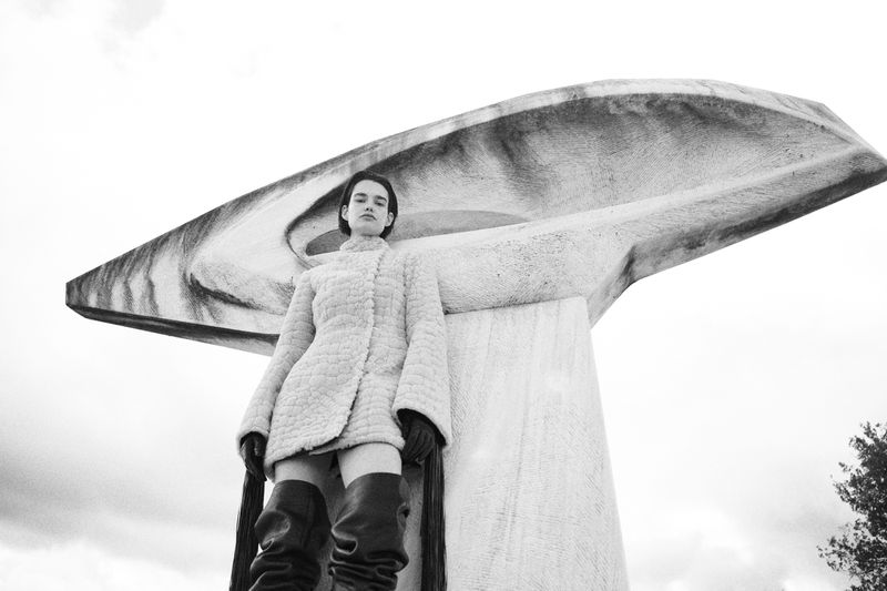 Woman in coat and boots stands in front of a concrete art structure against a cloudy sky in a black and white fashion shot.