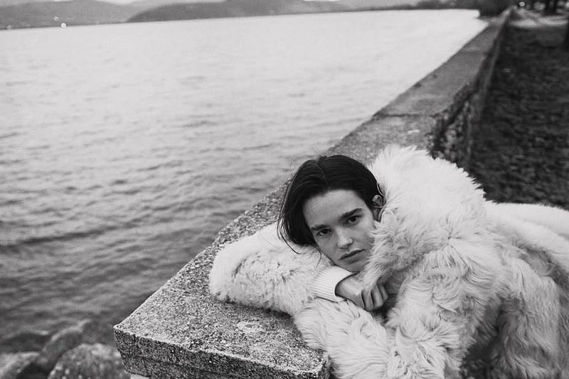 Monochrome portrait of a brunette female model in a white fur coat, lying on a stone ledge overlooking a lake in an outdoor setting.