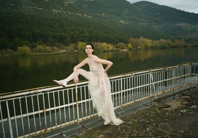 Woman in a sheer lace dress poses on a metal pier with a lakeside mountain backdrop. Fashion editorial photography, outdoor location.