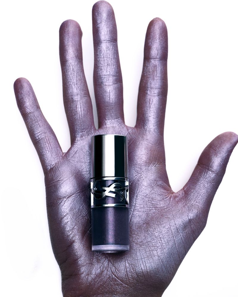 Hand with metallic purple paint holding a YSL lipstick tube against a white background in a beauty product shot.