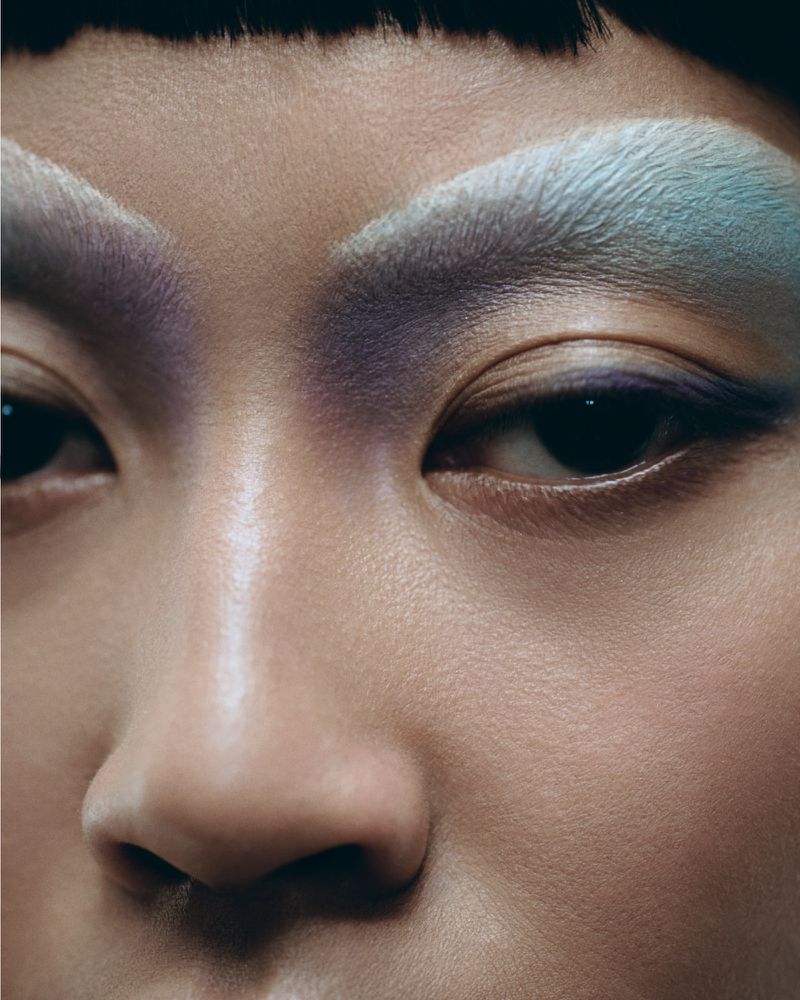 Close-up of an Asian woman's face with blue and purple eyeshadow. Her dark hair is visible at the top of the frame.