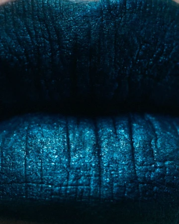 Close-up shot of a woman's lips covered in shimmering, metallic teal lipstick. The focus is on the texture and color of the makeup.