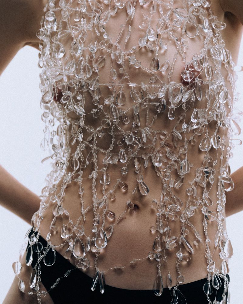Torso of an adult female wearing a top made of clear beads against a plain white backdrop in a studio. She is also wearing black bottoms.