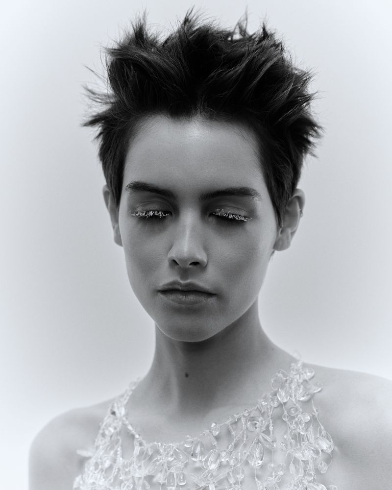 Black and white portrait of a young woman with short hair and glitter eyeliner, wearing a beaded top against a white background.