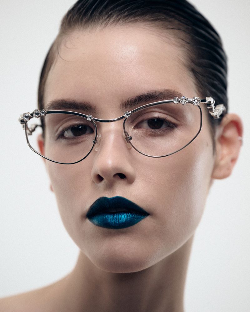 Close-up of a person with slicked-back hair and silver-rimmed glasses, featuring gems, and striking teal lipstick against a white backdrop.