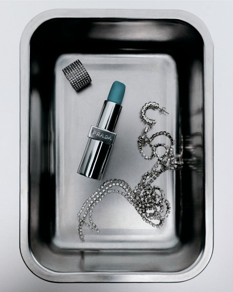 Still life of Prada lipstick, ring, earrings, and rhinestone necklace on a reflective metal surface, high-angle view, neutral background.