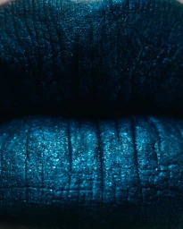 Extreme macro close-up of lips covered in textured shimmering dark teal metallic lipstick against a dark frame.