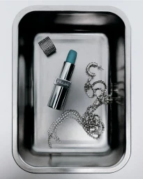 Silver lipstick with teal blue bullet, sparkling rhinestone chain, and ring inside a stainless steel metal tray on a white surface.