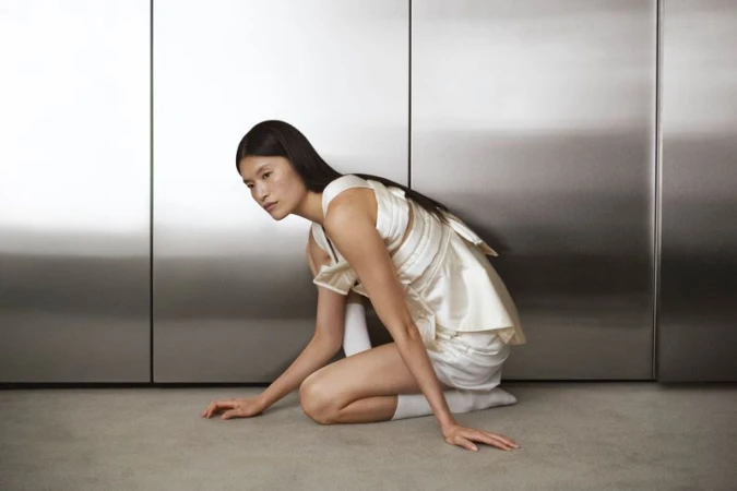 Asian woman in a white satin dress poses kneeling indoors against a modern metal wall, with soft lighting and a serene expression.