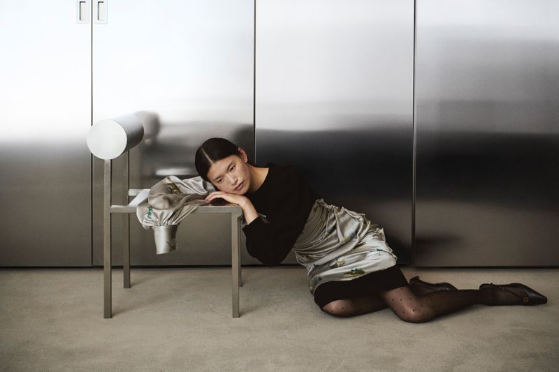 Young Asian woman in black top and patterned skirt lies next to a sculptural silver chair against a metallic silver panel backdrop.