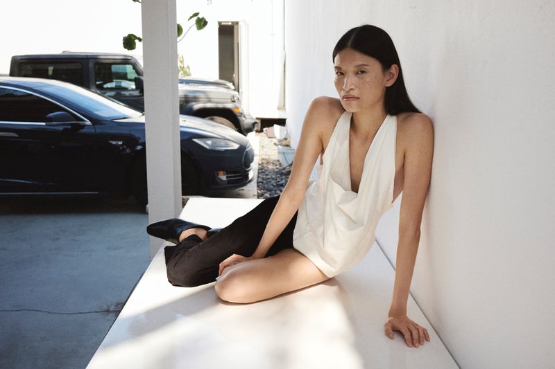 Elegant Asian woman in halter top and dark pants poses in a modern outdoor space with luxury cars in the background on a sunny day.