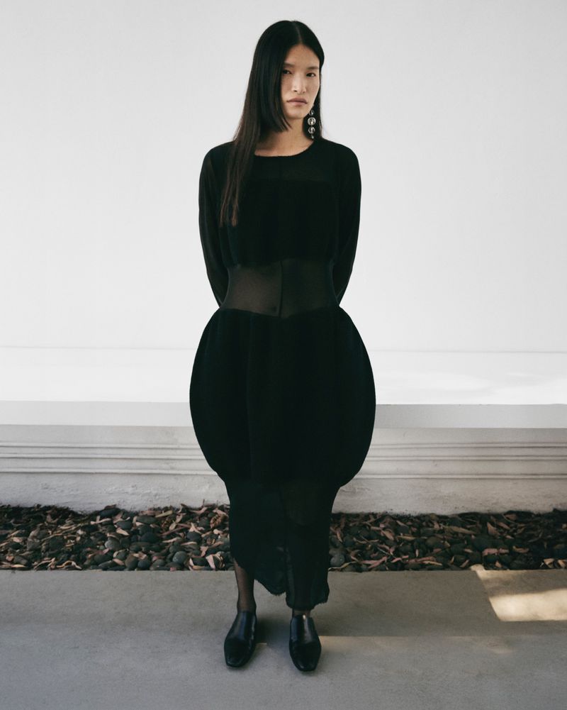 Full-length shot of a stylish woman in a black dress with a wide belt, complemented by black shoes, against a neutral background.