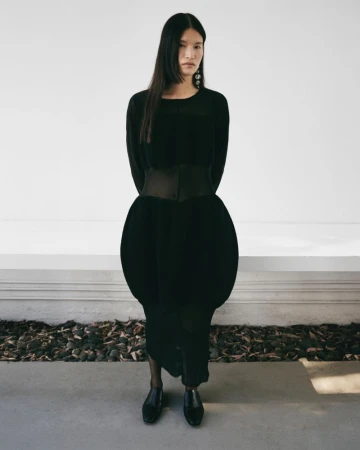 Full-length shot of a stylish woman in a black dress with a wide belt, complemented by black shoes, against a neutral background.