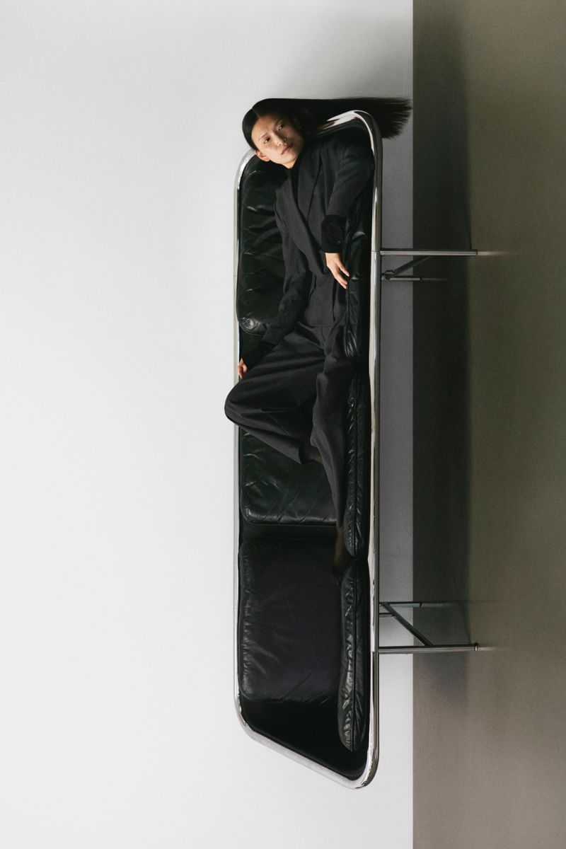 Elegant Asian woman in black suit lying on black leather sofa, indoor setting against contrasting white and gray walls in a portrait orientation.