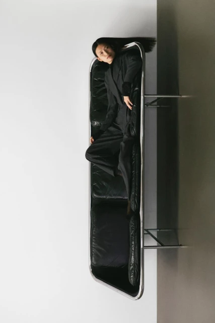 Elegant Asian woman in black suit lying on black leather sofa, indoor setting against contrasting white and gray walls in a portrait orientation.