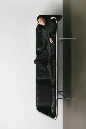 Elegant Asian woman in black suit lying on black leather sofa, indoor setting against contrasting white and gray walls in a portrait orientation.